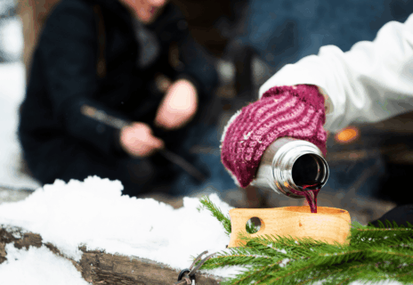 Talvinen kuva laavulta, jossa naisen käsi lapasessa ja käsi kaataa termarista juomaa.