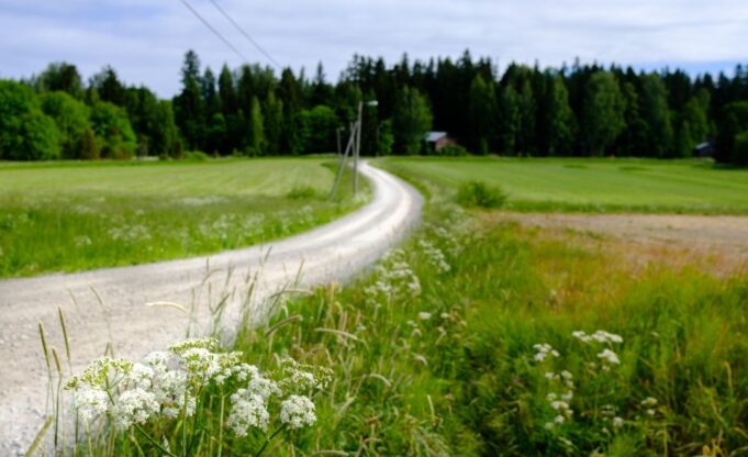 Soratie kiemurtelee vihreiden peltojen läpi, taustalla metsä ja pieni rakennus.