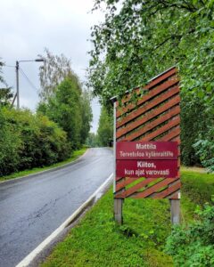 Kuvassa on kylänraitti, jonka varrella on kyltti. Kyltissä lukee "Mattila – Tervetuloa kylänraitille. Kiitos, kun ajat varovasti." Kyltin tausta on luonnonläheinen, ja ympäristö vaikuttaa rauhalliselta ja maaseutumaiselta.