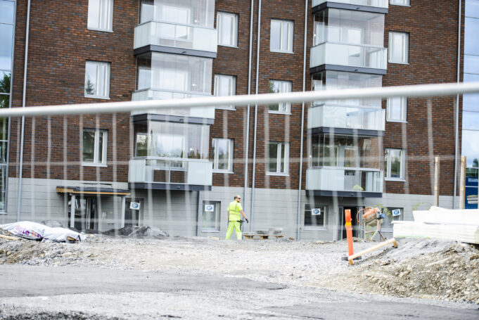 Taustalla rakenteilla oleva ruskea kerrostalo, jonka edessä huomiovaatetteissa rakennustyöntekijä. Kuvanedessä verkkoaita.