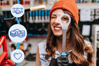 Pitkähiuksinen nuori henkilö kurkistaa toisella silmällä ison kiintoavaimen reiän läpi. Taustalla pyöränkorjauspaja työkaluineen.