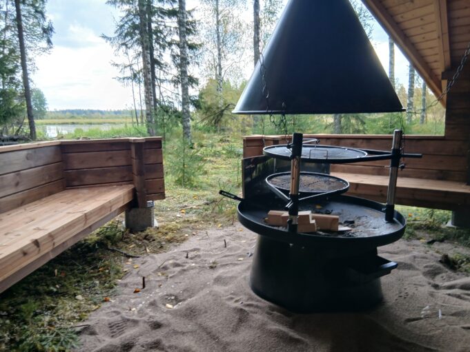 Näkymä kodasta vesistöä kohti. Etualalla tulipaikka ja kodan penkkejä, taustalla näkyy rantametsää, vesistöä ja sen takana ruovikkoa.